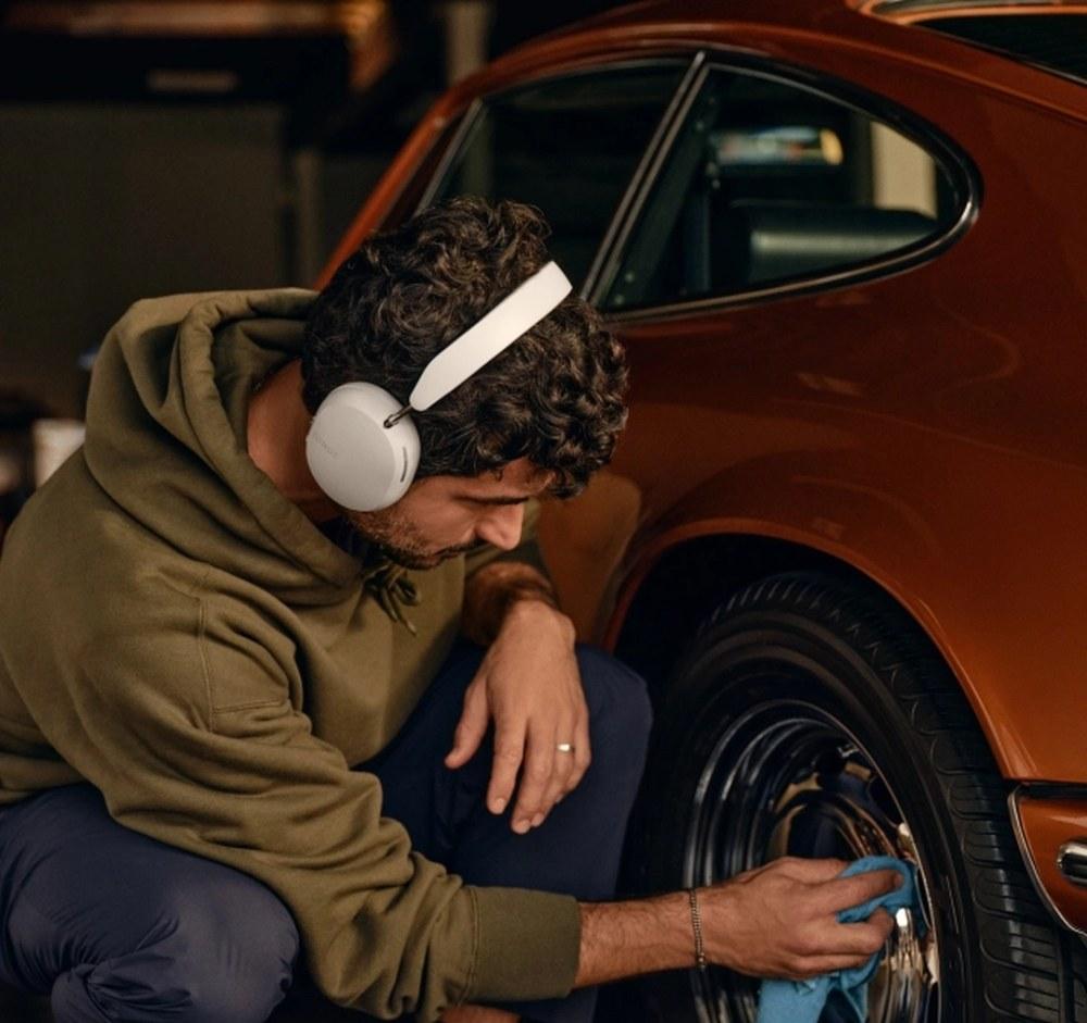 Férfi felhasználó egy pár puha fehér Sonos Ace fejhallgatót visel, miközben egy klasszikus autó kerekeit fényezi.